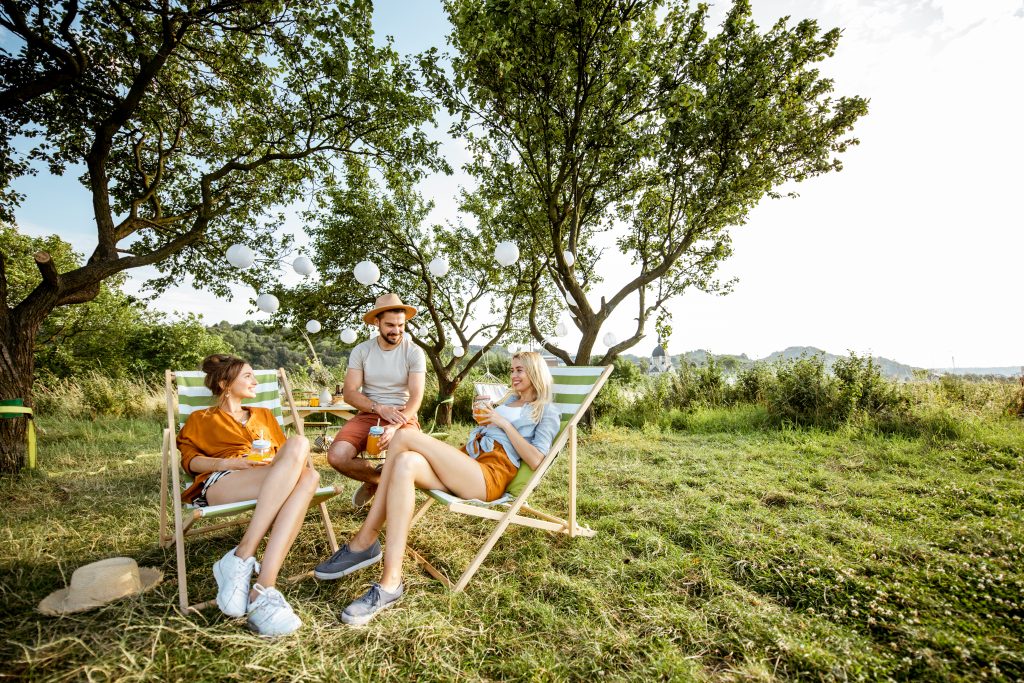 Young and cheerful friends relaxing with drinks, sitting on the sunbeds in the beautifully decorated backyard, wide view with copy space